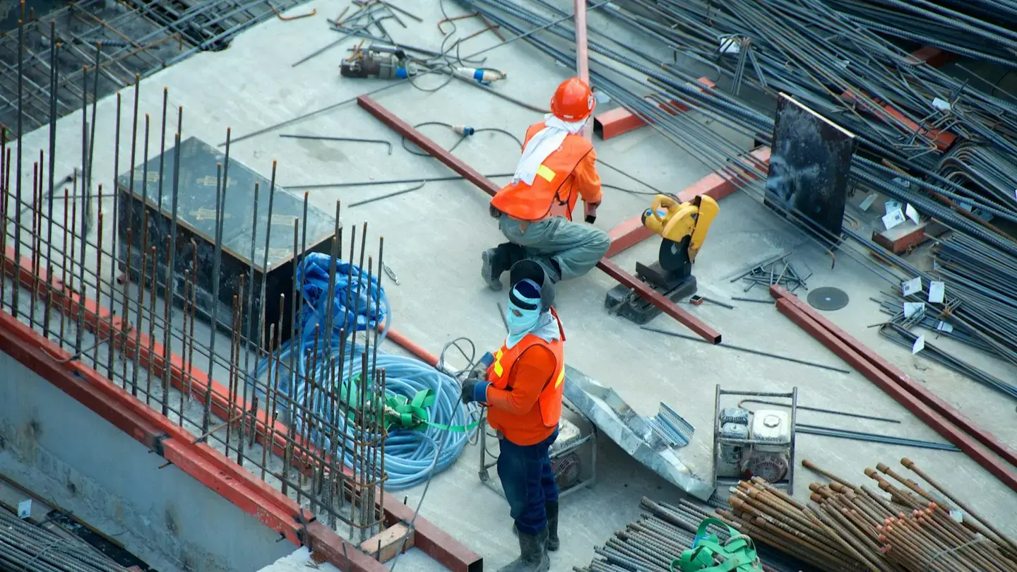 Ouvriers en gilet orange et casque travaillant sur une dalle de chantier encombrée d’armatures
