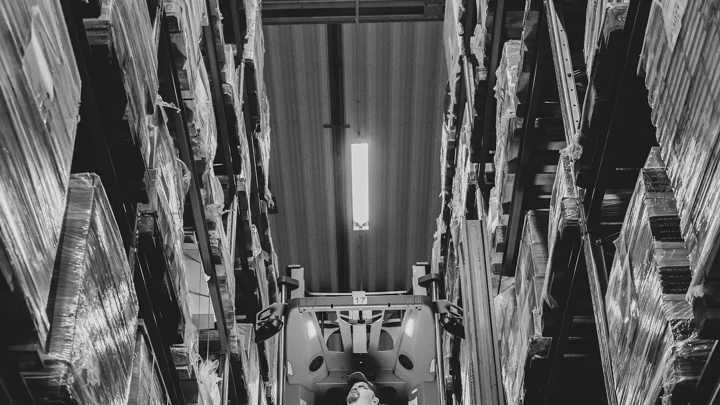 Vue en noir et blanc d’un chariot élévateur dans une allée étroite entre de hauts rayonnages de palettes filmées