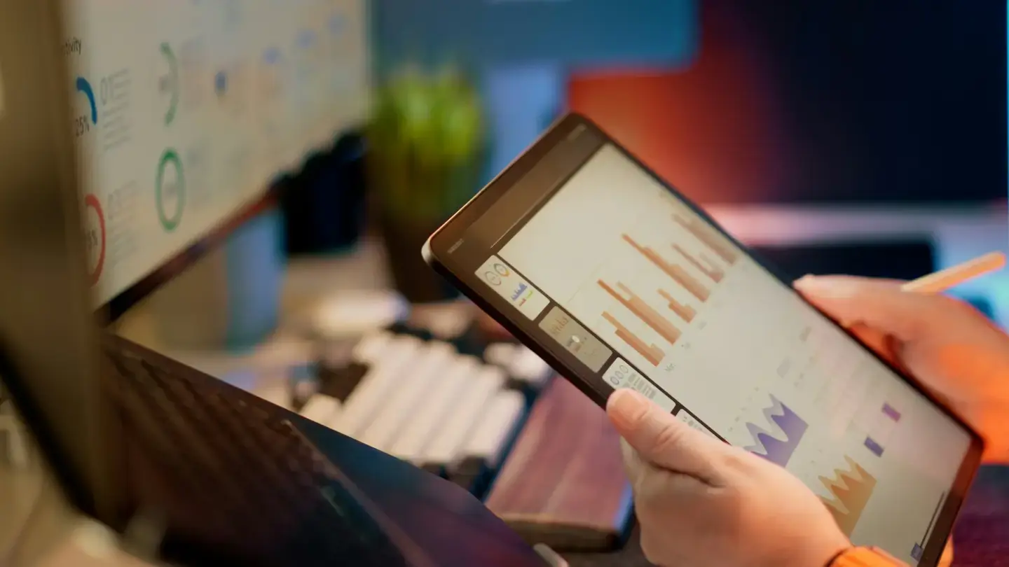 Une personne consulte des graphiques en barres sur une tablette devant un écran d’ordinateur