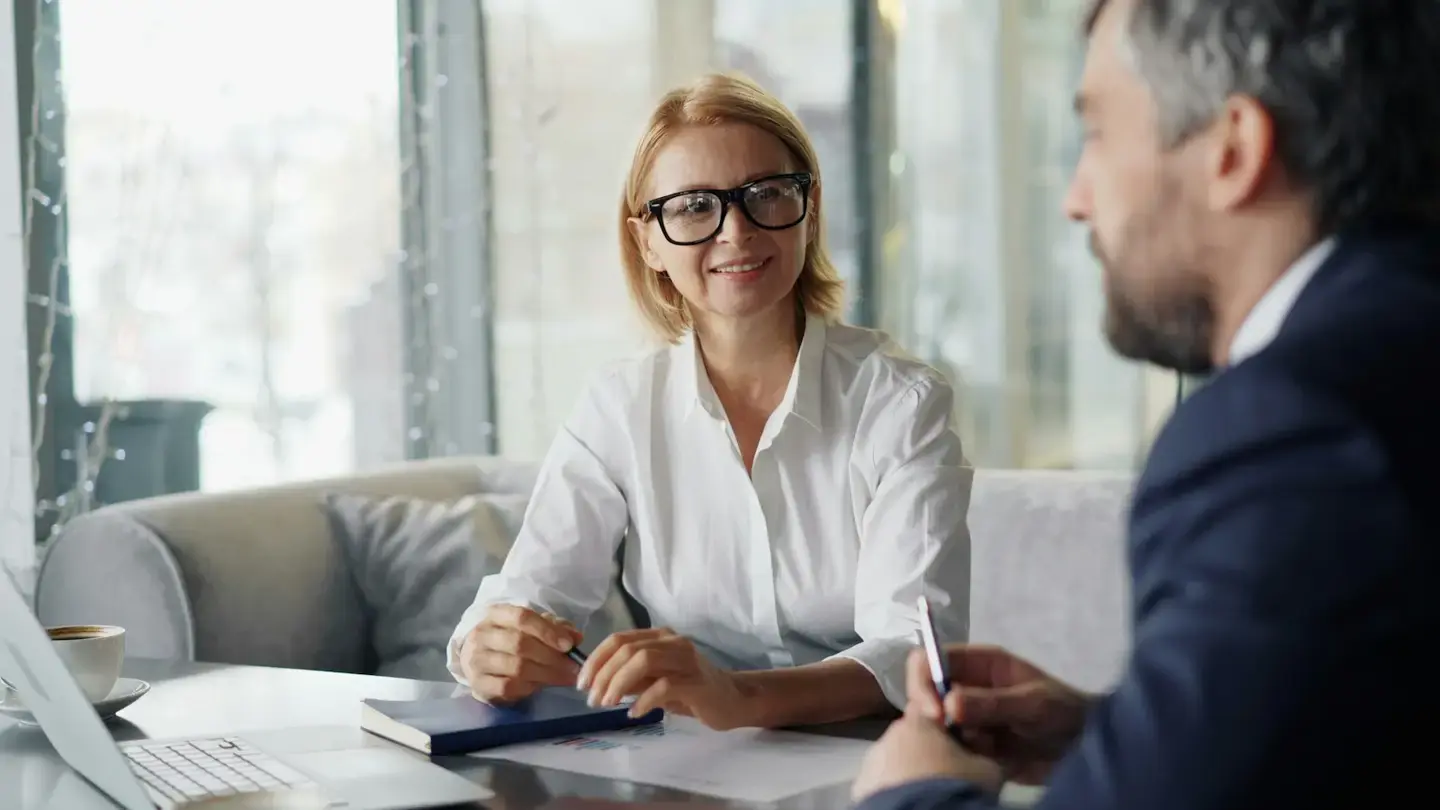 Une commerciale échange avec un client autour d’une table, ordinateur portable et documents posés devant eux