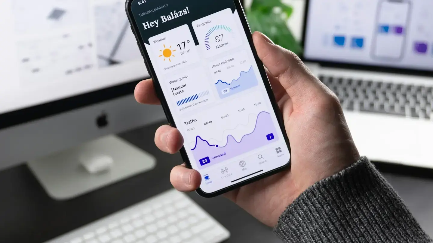 Une main tient un smartphone affichant un tableau de bord, devant un ordinateur de bureau et un portable