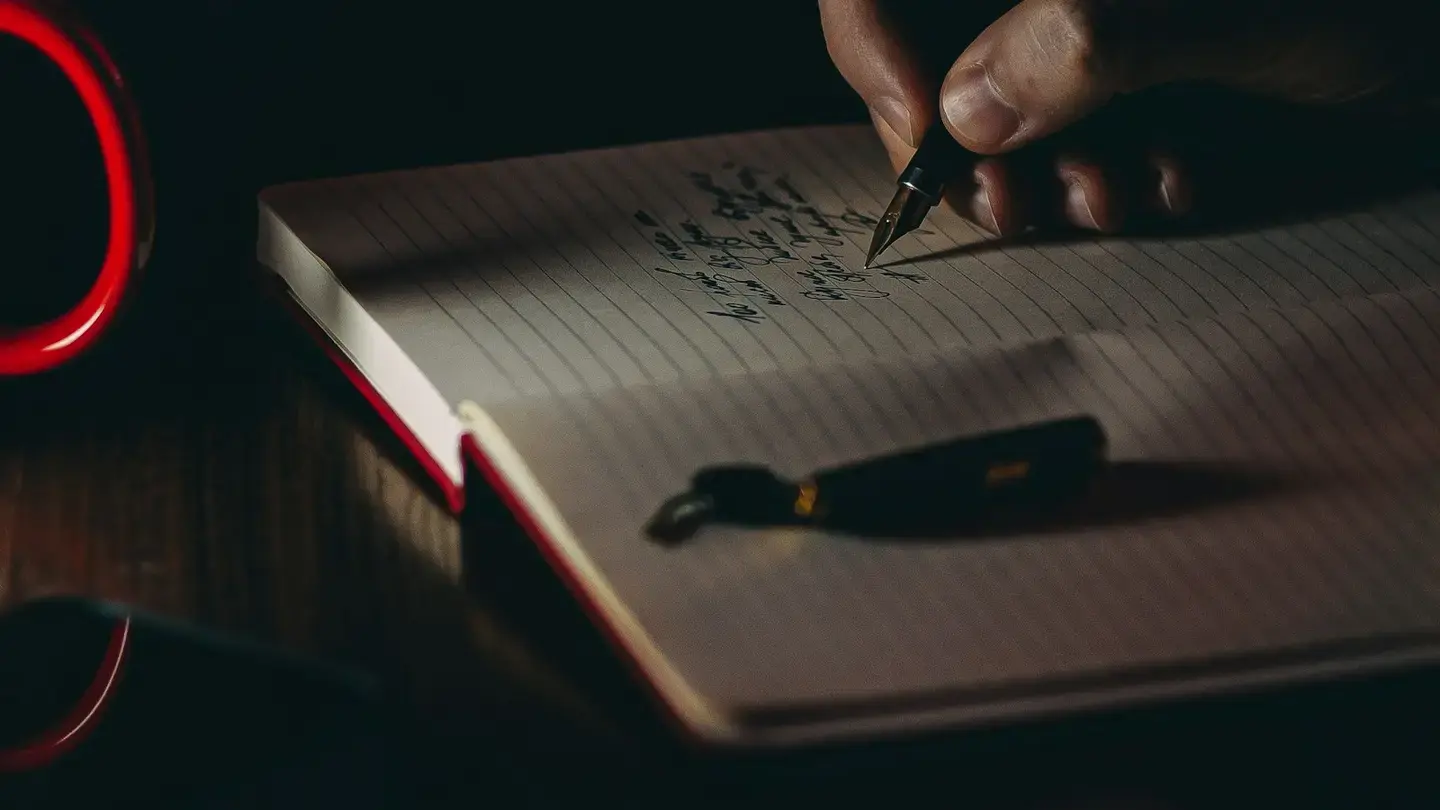 Main qui écrit à l’encre dans un carnet ouvert posé sur un bureau sombre