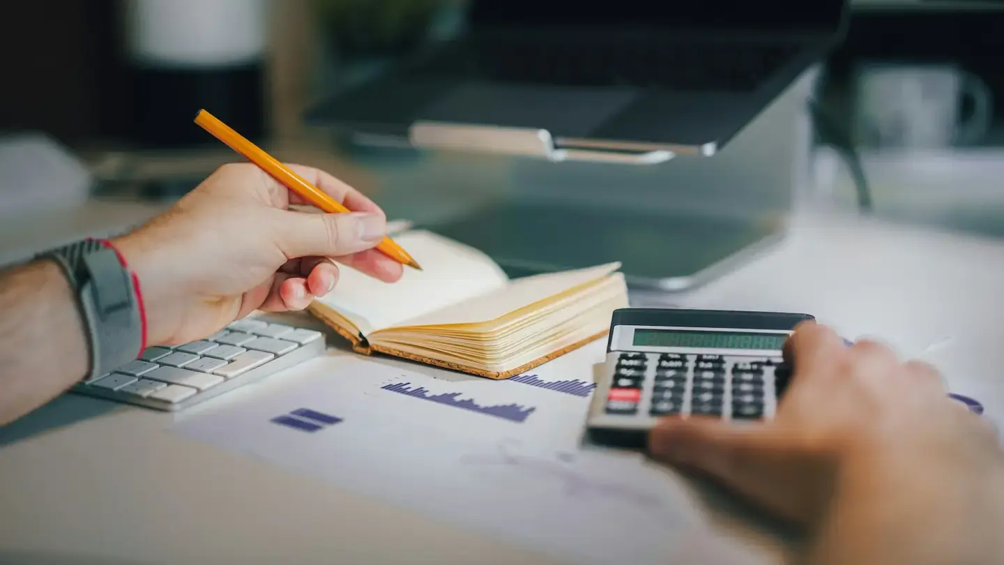 Mains tenant un crayon au-dessus d’un carnet et utilisant une calculatrice posée sur des graphiques imprimés