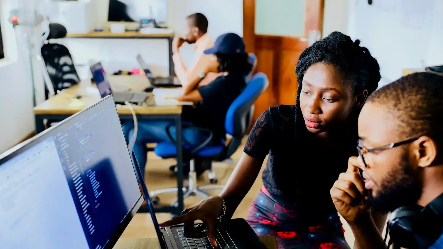Deux développeurs examinent ensemble du code sur un écran, des collègues travaillent en arrière-plan