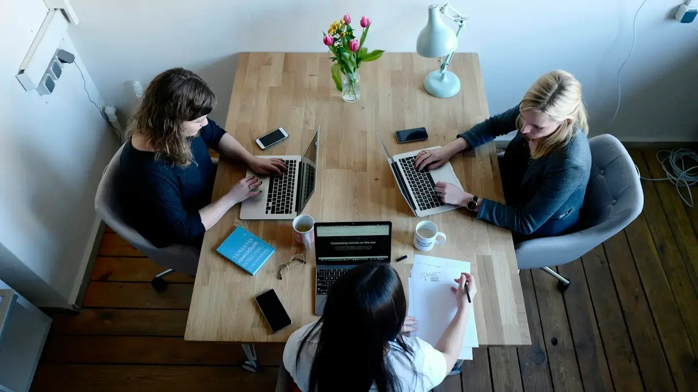 Trois collaboratrices travaillent autour d’une table en bois avec ordinateurs portables et documents