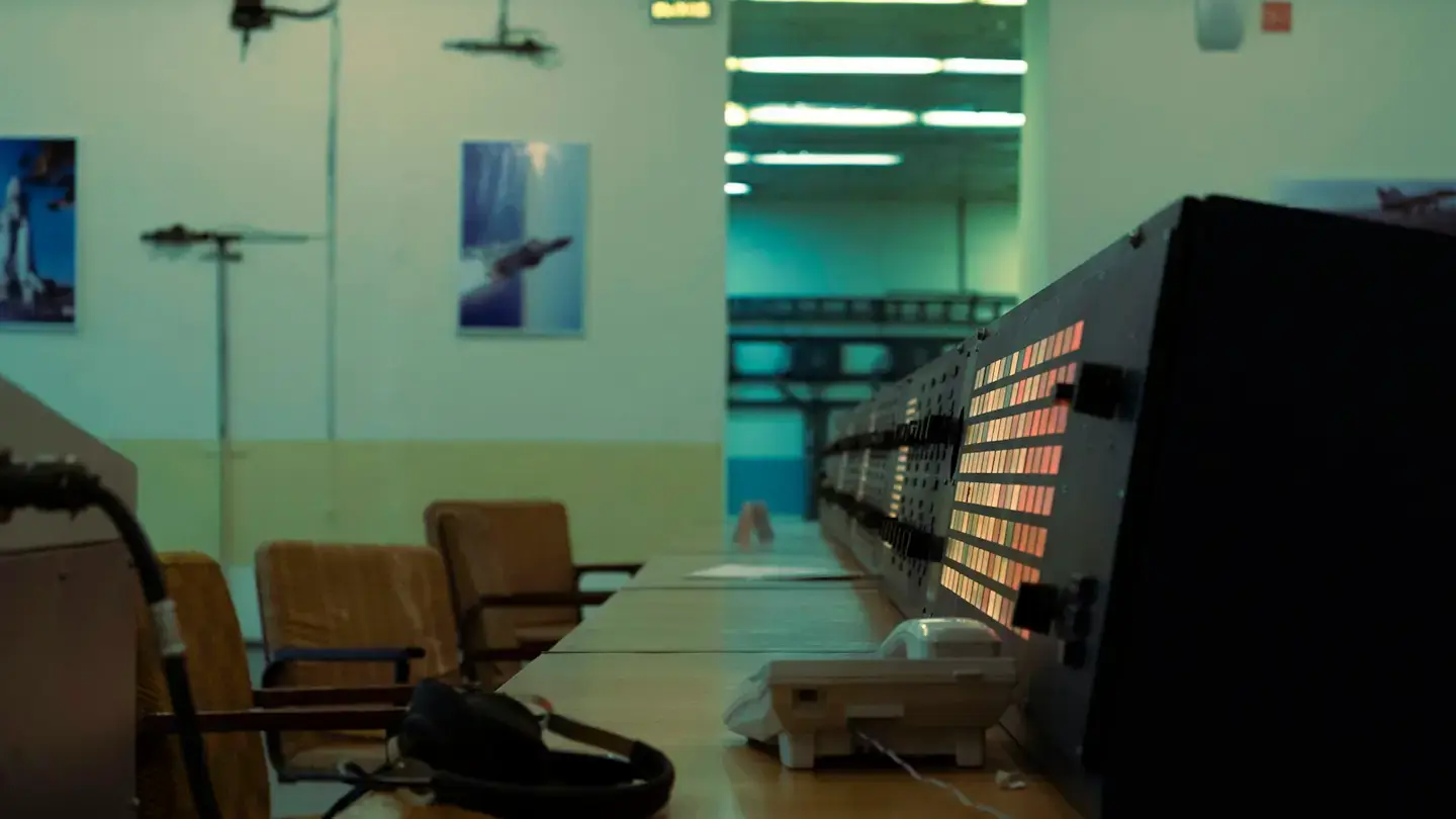 Salle de contrôle avec un long pupitre de voyants lumineux, un téléphone et un casque posés sur le bureau
